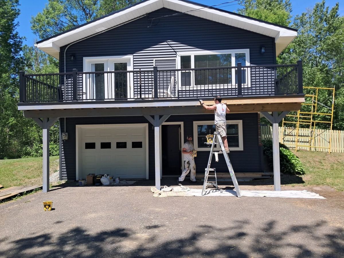 Two-story navy blue home with white garage and balcony, Freedom Painting crew on ladder finishing the eaves
